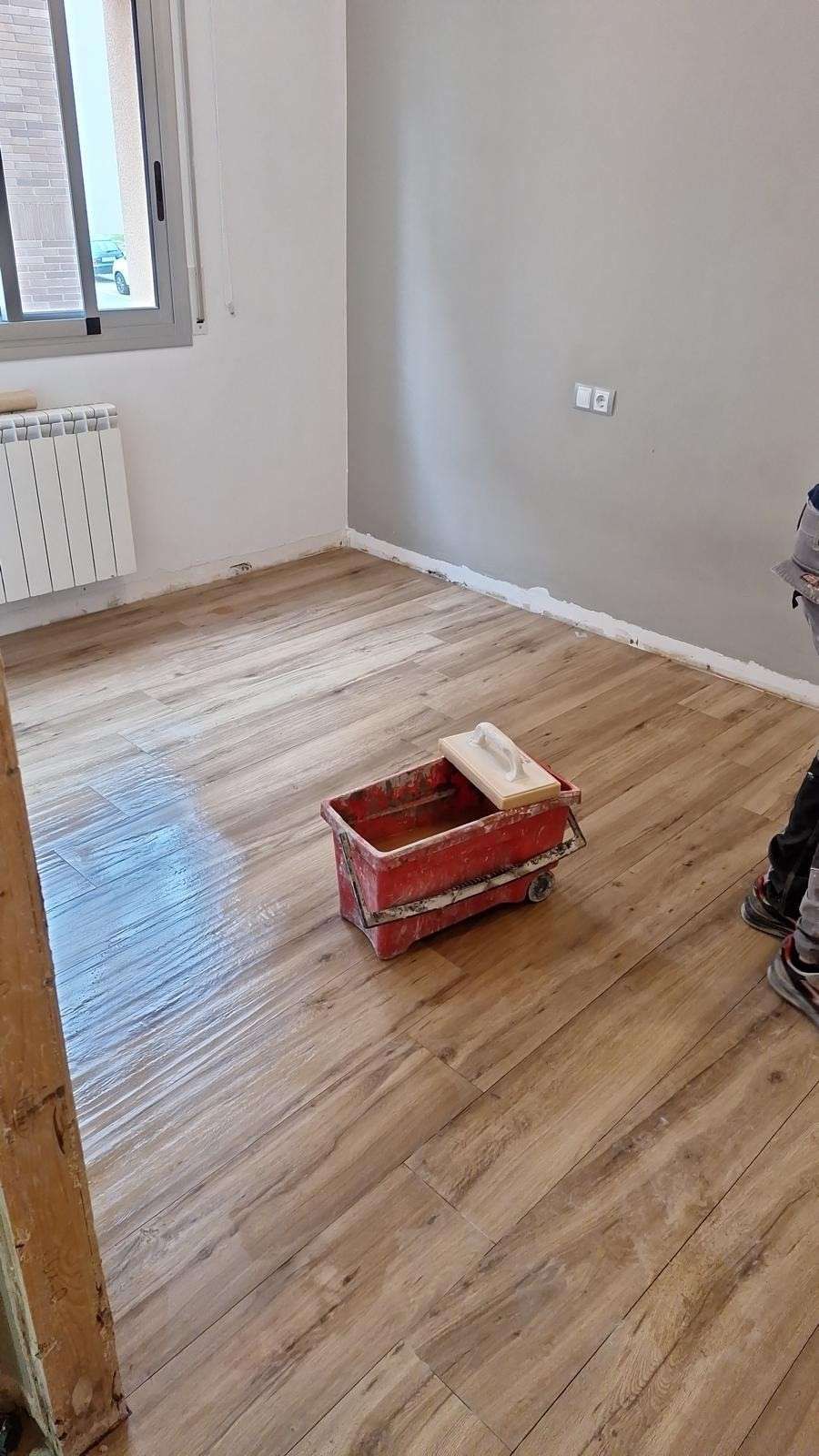 Habitación vacía con suelo que imita la madera, ventana, radiador y una caja roja en el suelo.