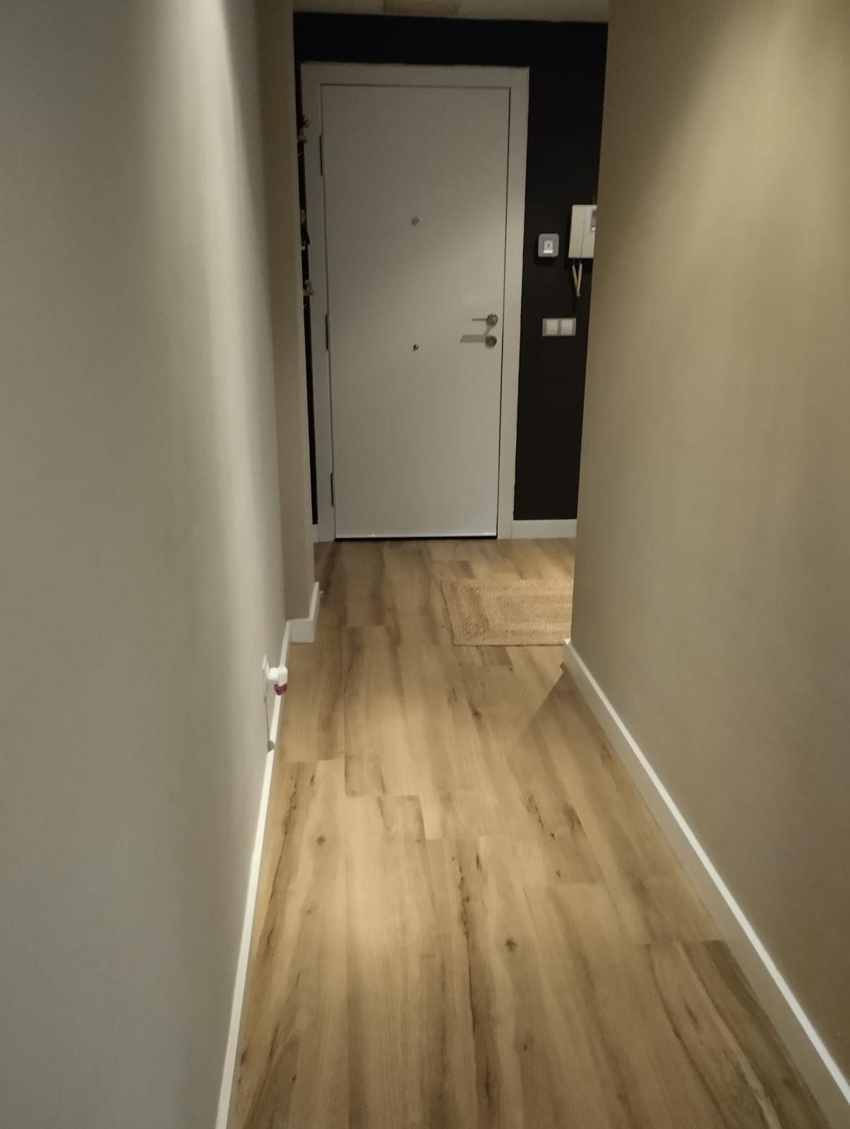 Pasillo estrecho con suelo de madera, paredes de color beige y una puerta blanca al final.