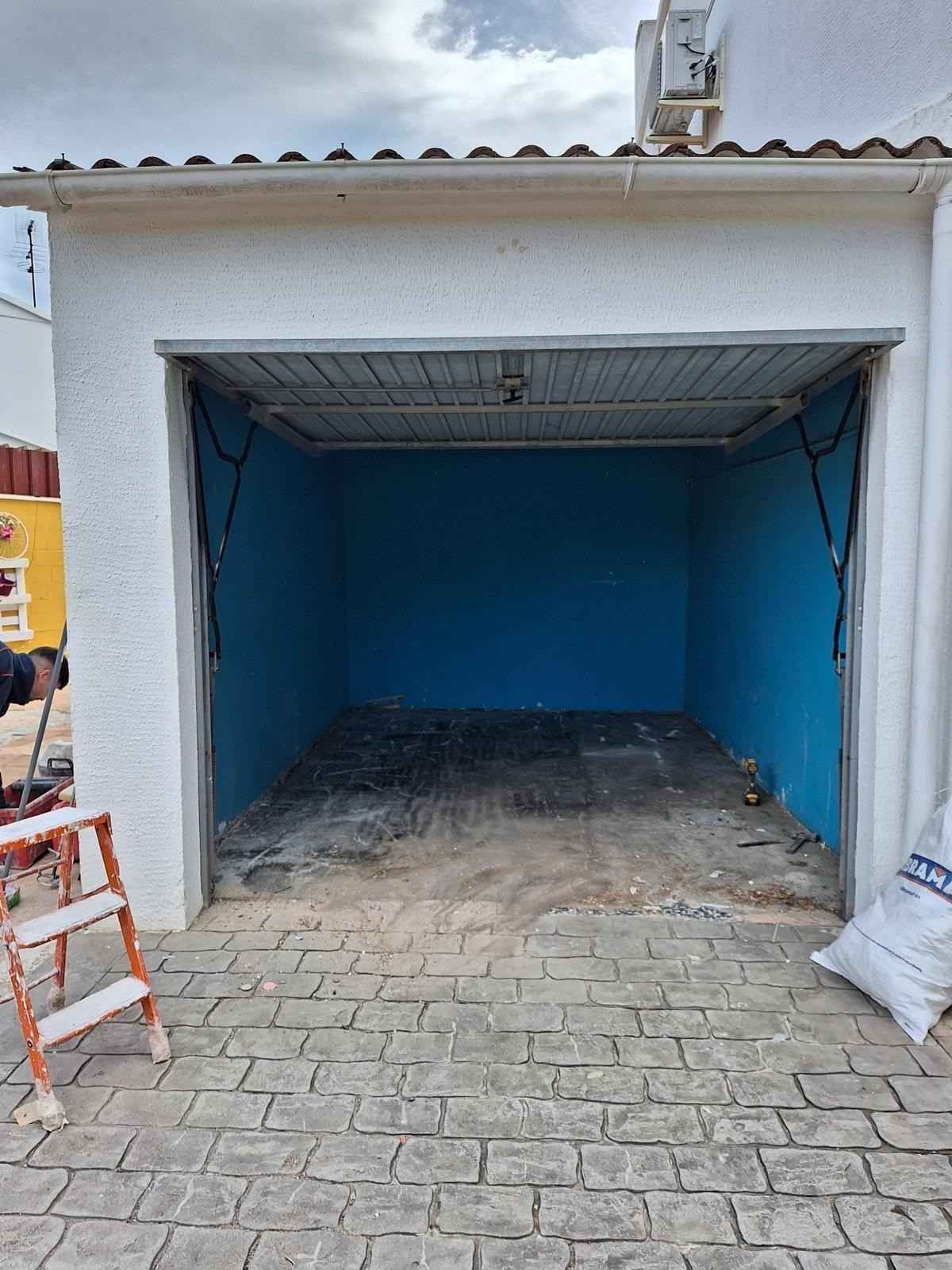 Garaje azul vacío con puerta metálica abierta y entrada pavimentada con piedra.