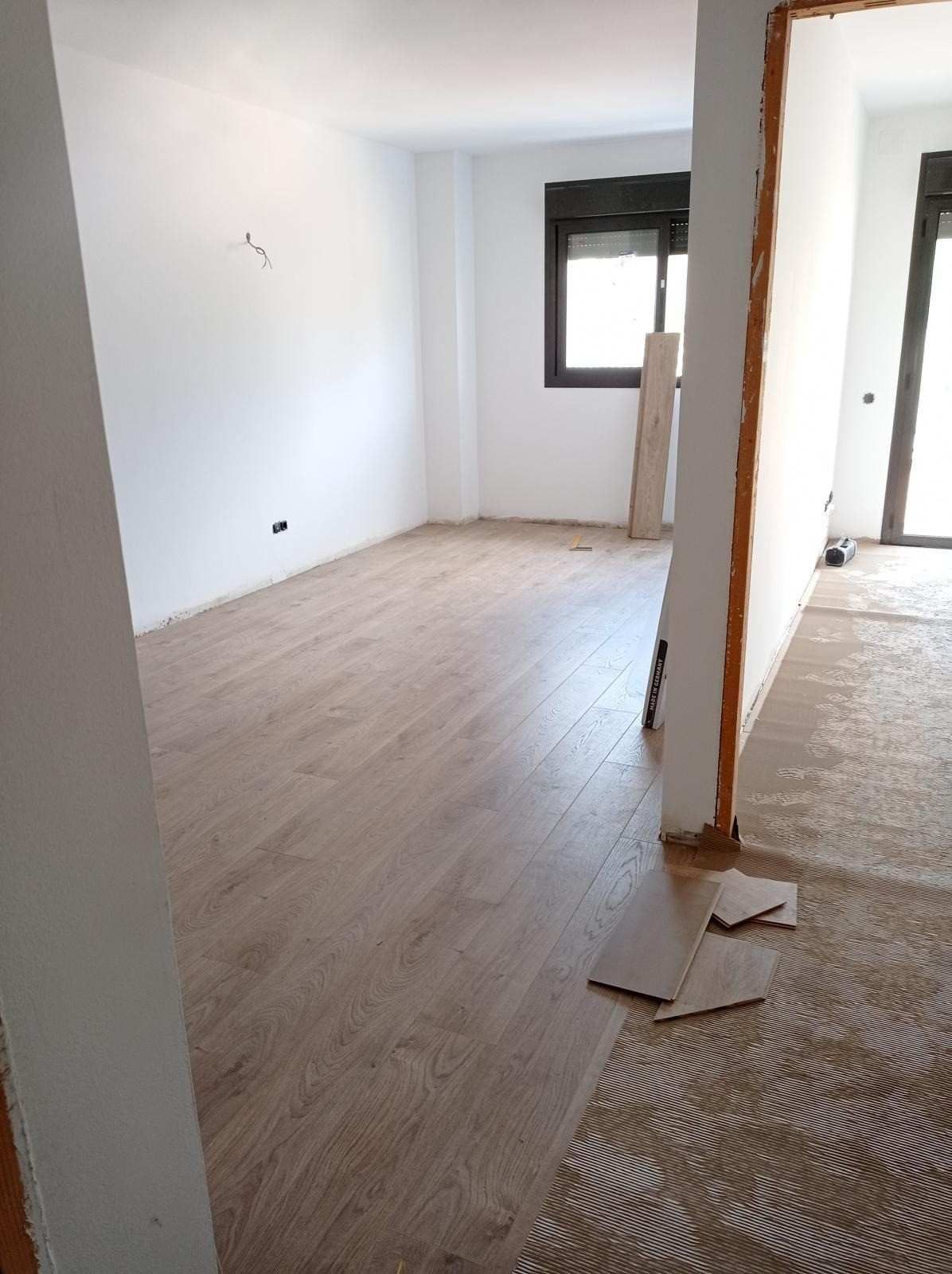 Habitación vacía con suelo de madera clara, paredes blancas y una ventana, parcialmente en obras.