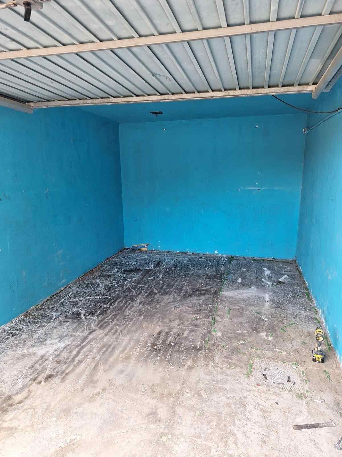 Caseta de almacenamiento azul vacía con techo de chapa ondulada y suelo de hormigón desgastado.