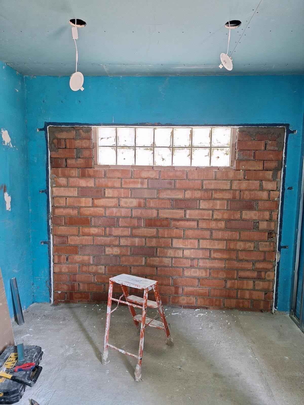 Habitación sin terminar con paredes azules, pared trasera de ladrillo, luces de techo a la vista y un pequeño taburete sobre un suelo de hormigón.
