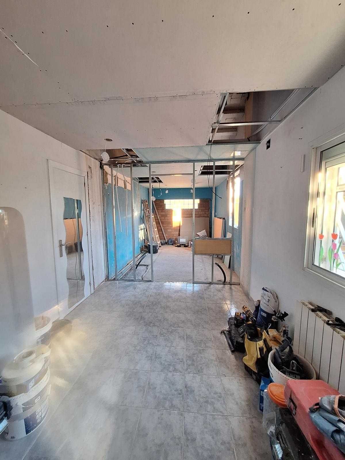 Habitación sin terminar con paredes desnudas, vigas de techo a la vista, escaleras y herramientas en proceso de renovación.
