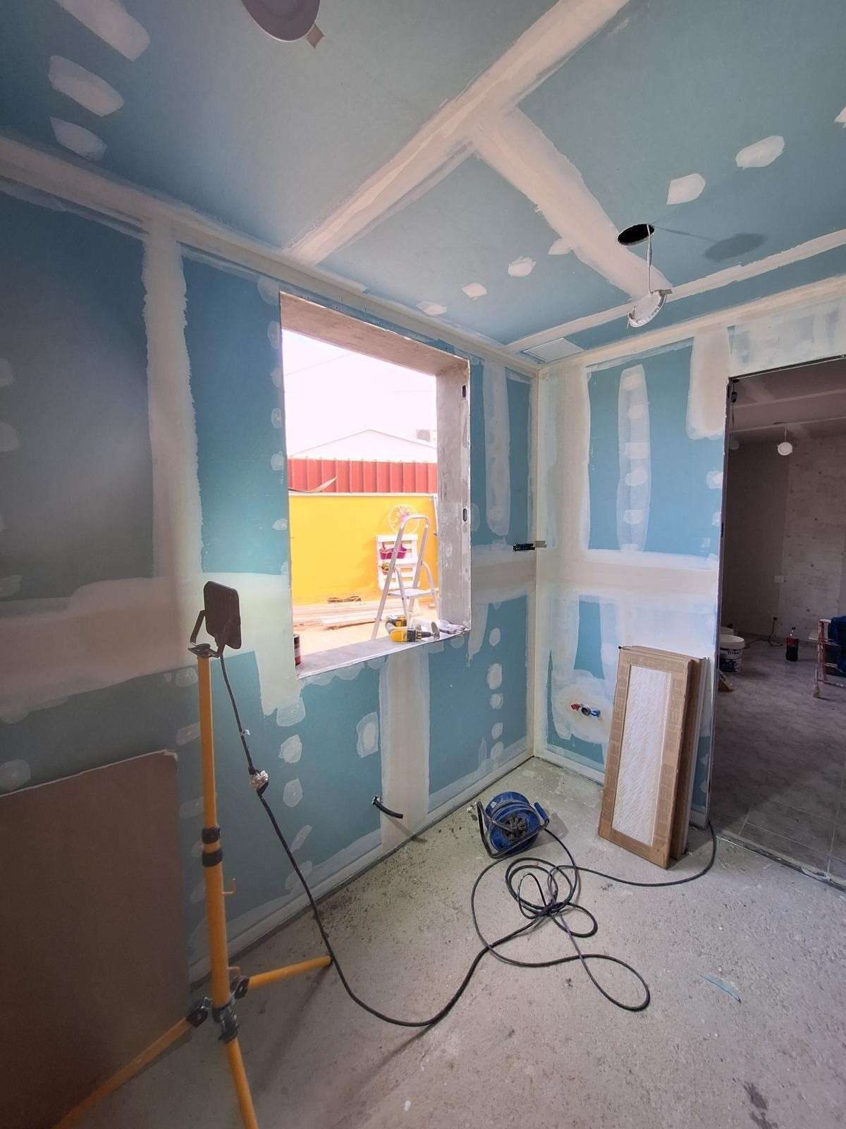 Habitación parcialmente reformada con paredes de pladur azul, juntas a la vista, una ventana, herramientas y cables en el suelo.