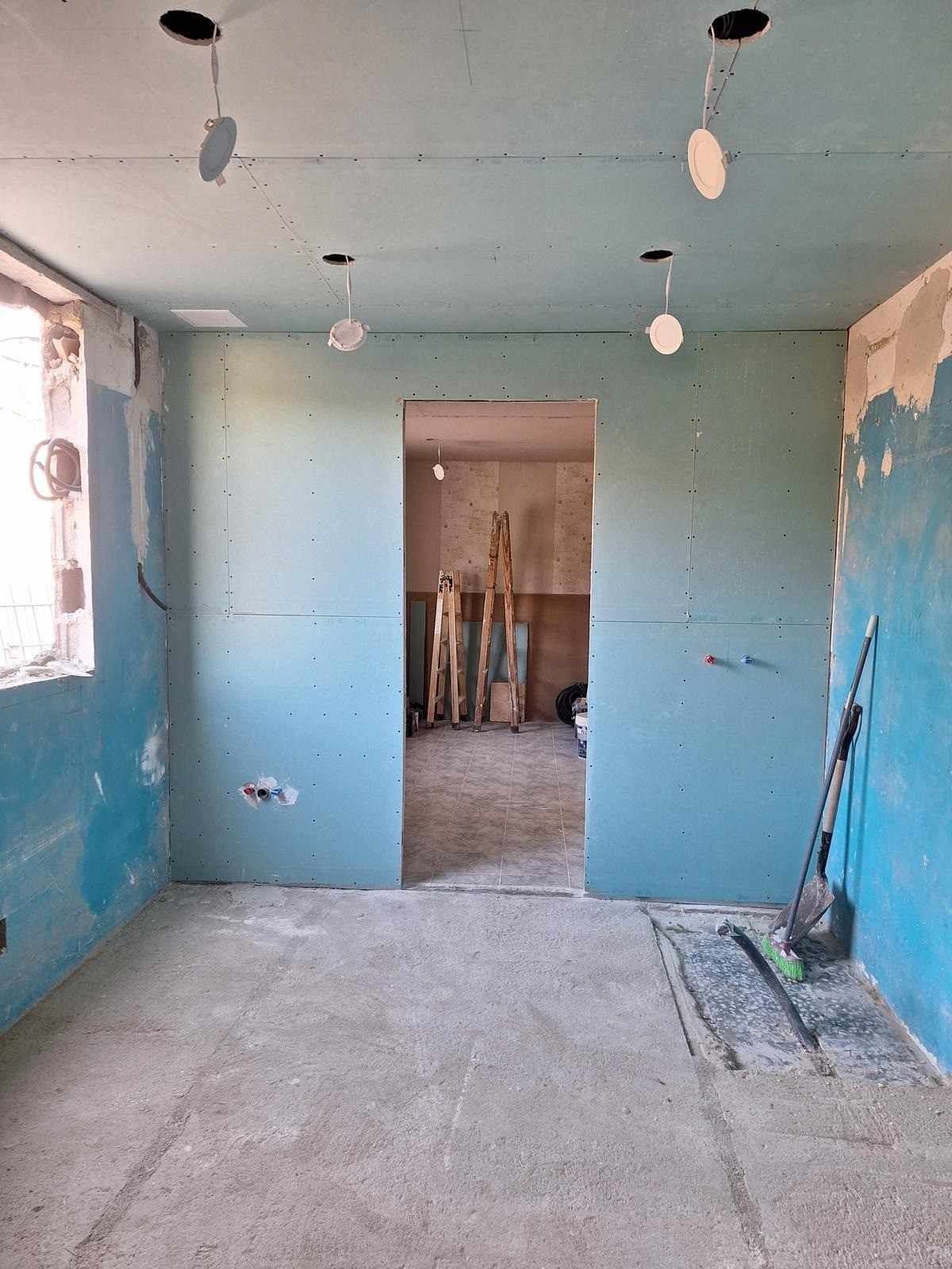 Habitación vacía en obras con paredes azules, luces de techo sin terminar y una puerta que da a otra habitación.