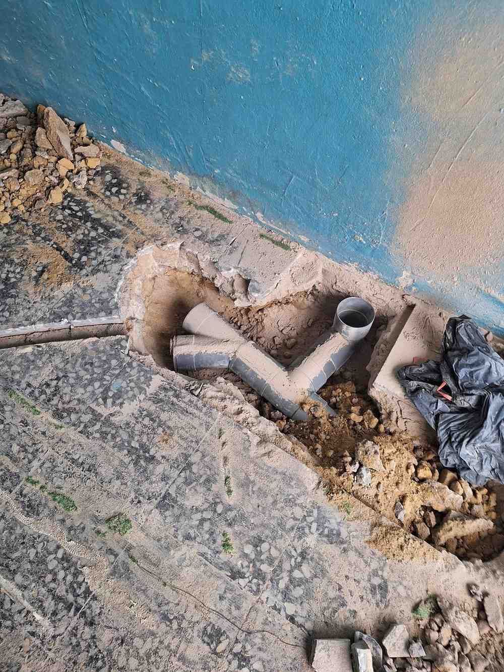 Zanja excavada junto a una pared azul con tuberías expuestas y escombros en el suelo.