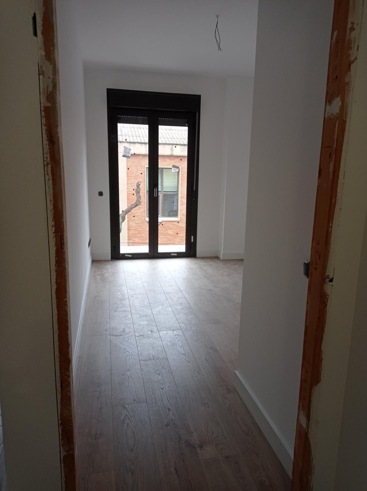 Pasillo estrecho con paredes blancas y suelo de madera que conduce a unas puertas de cristal que dan a un balcón.