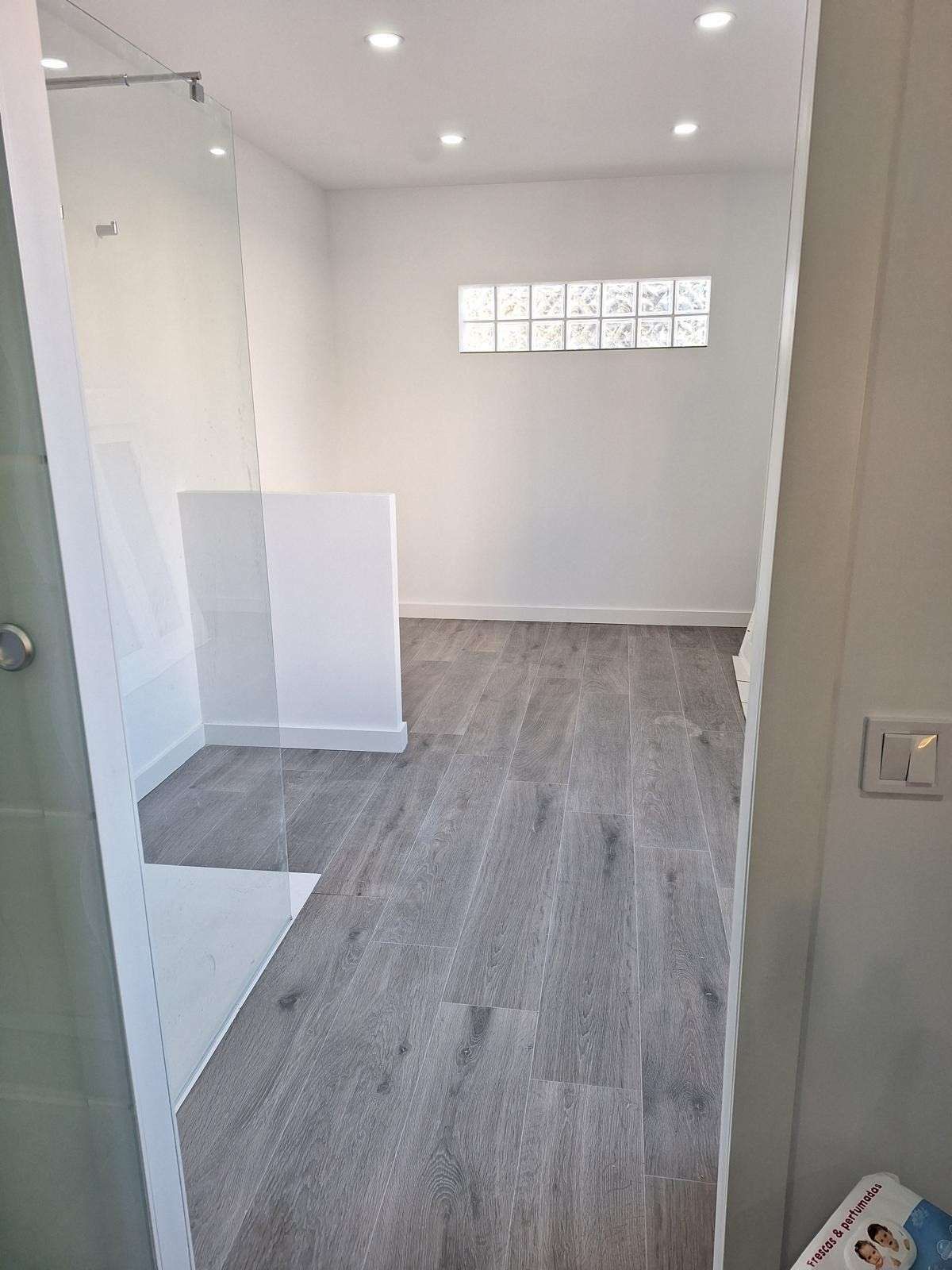 Habitación moderna y vacía con suelos de madera gris, paredes blancas, ducha de cristal y pequeñas luces esmeriladas en las ventanas.