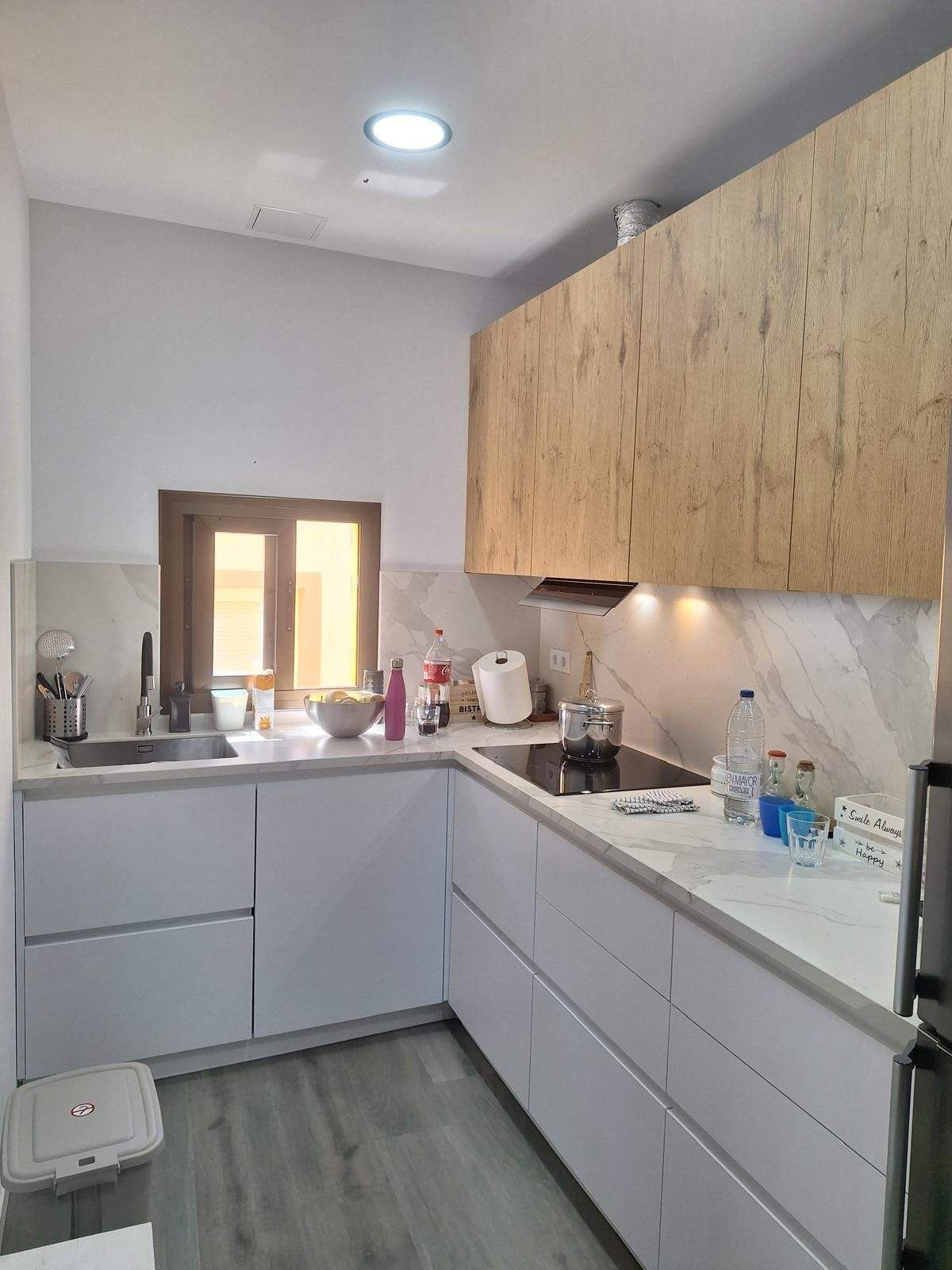 Cocina moderna y compacta con gabinetes gris claro, gabinetes superiores de madera, encimera blanca y fregadero pequeño debajo de una ventana.