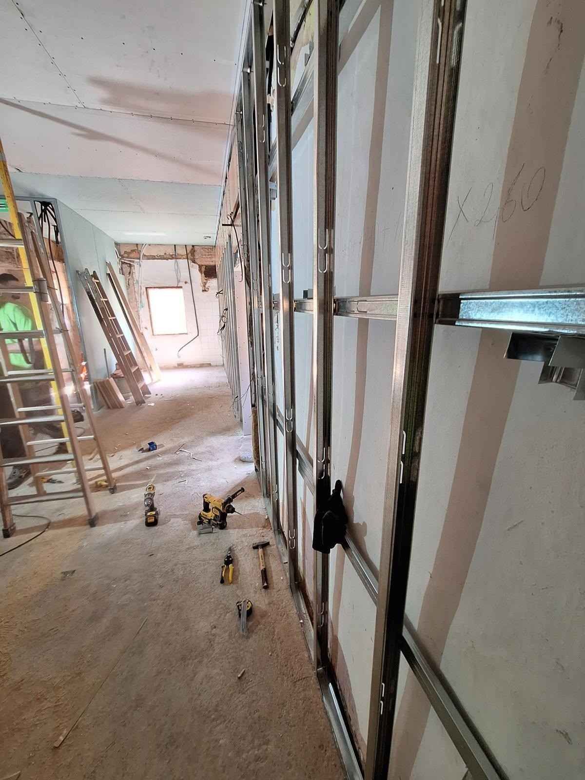 Pasillo estrecho en obras con estructura a la vista, herramientas en el suelo y escaleras al fondo.