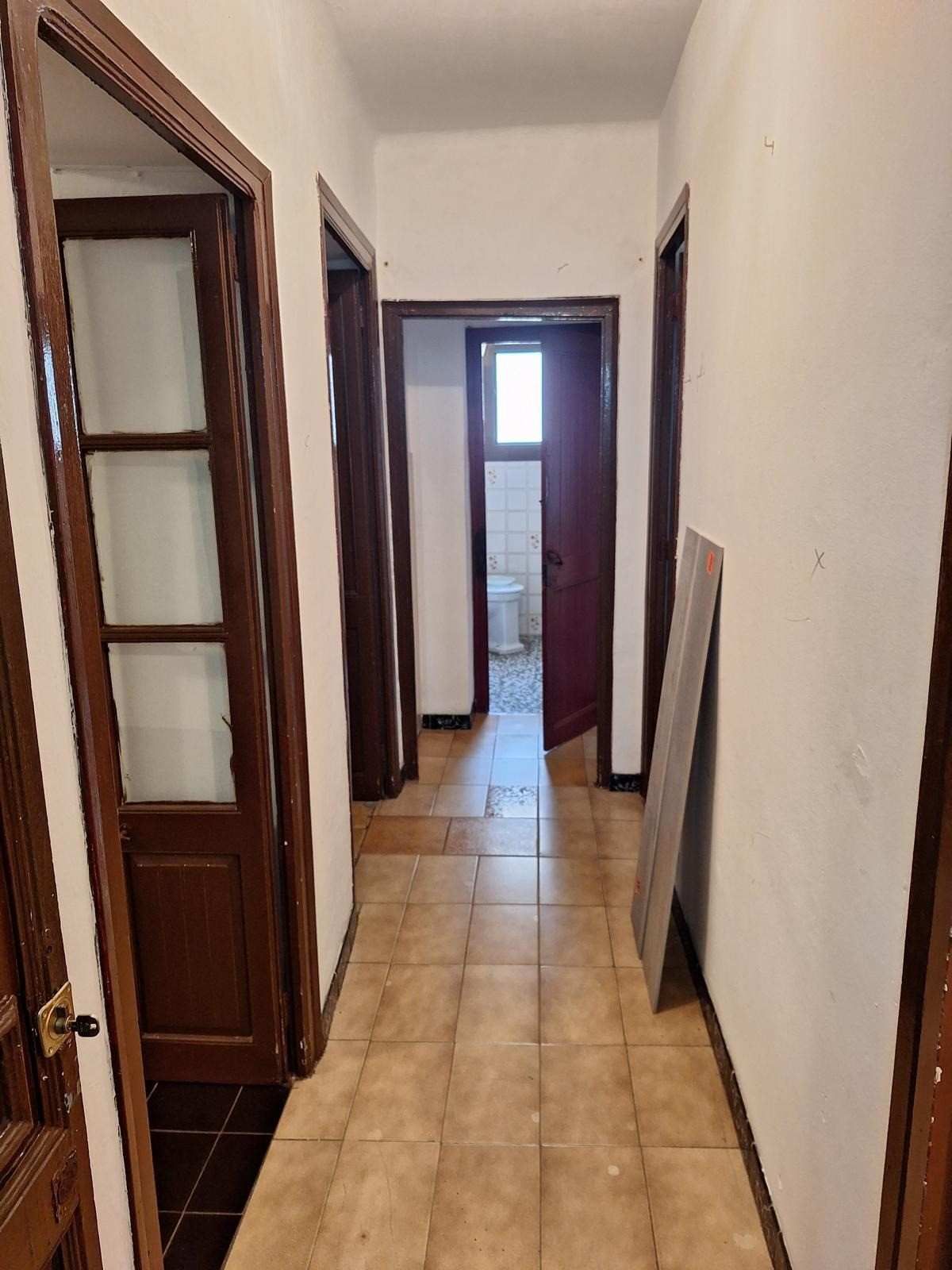 Pasillo estrecho con suelo de baldosas, paredes blancas y puertas de madera oscura, que conduce a una puerta luminosa al final.