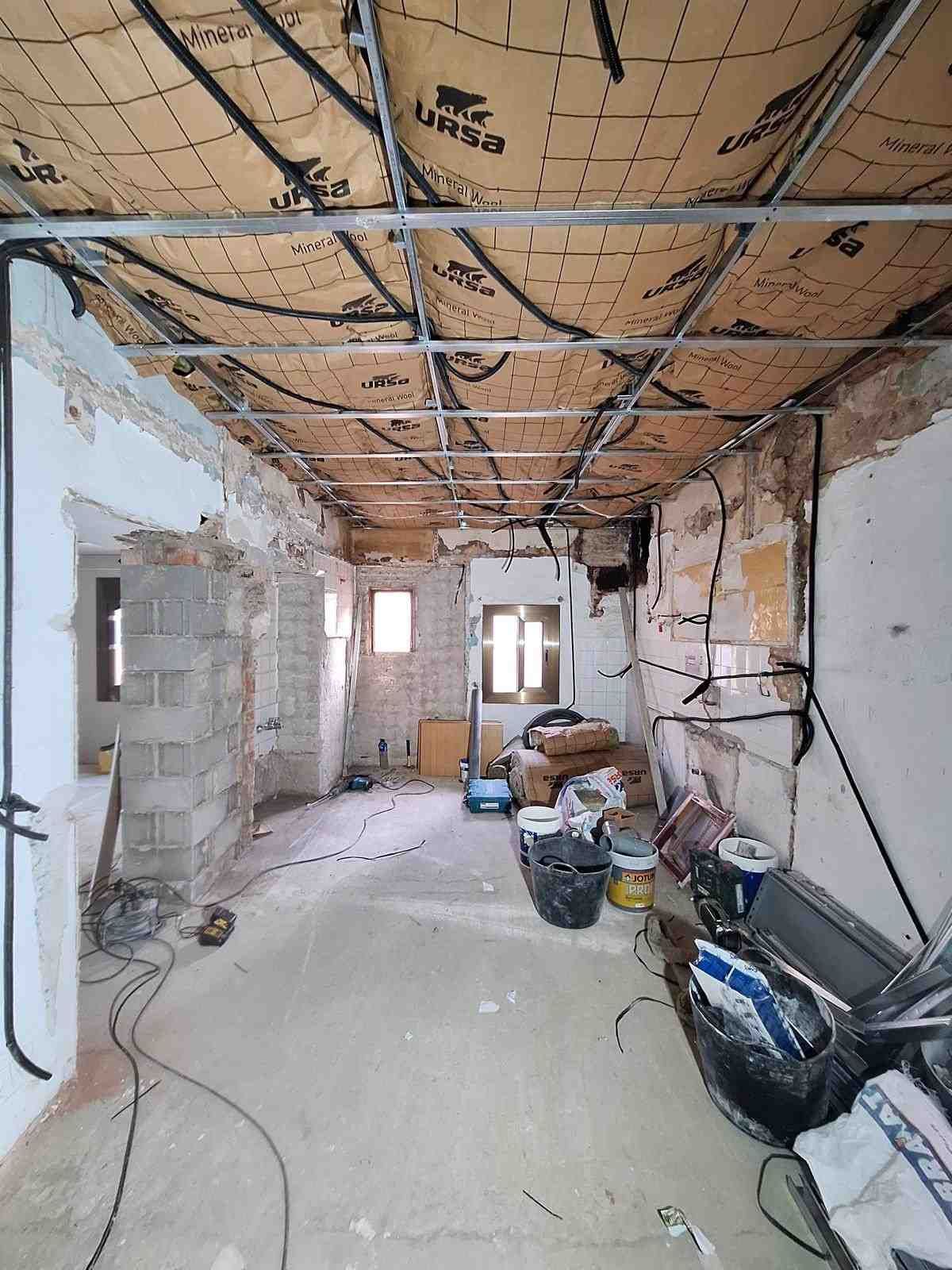 Obras de renovación en curso en una habitación estrecha, con el aislamiento del techo a la vista, el cableado y herramientas en el suelo.