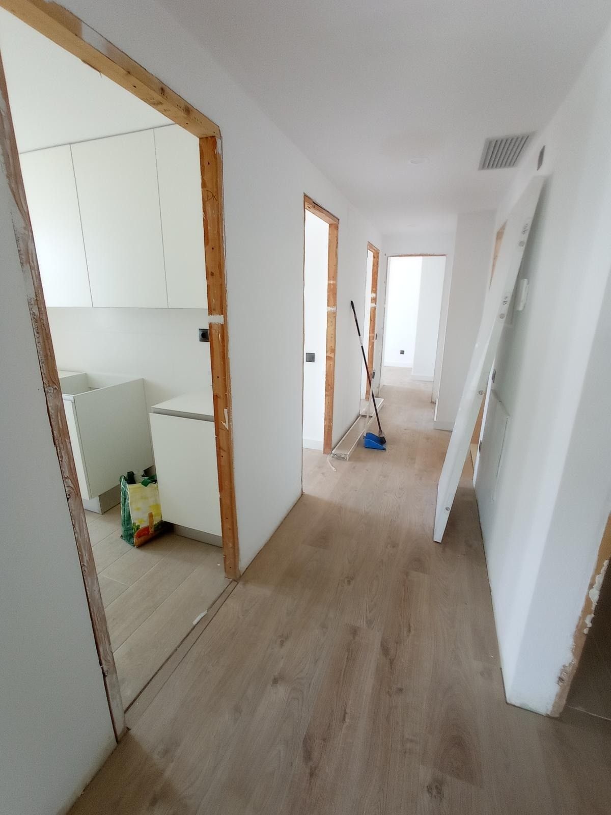Pasillo vacío en una casa con paredes blancas, suelos de madera clara y una puerta que da a un baño a la izquierda.
