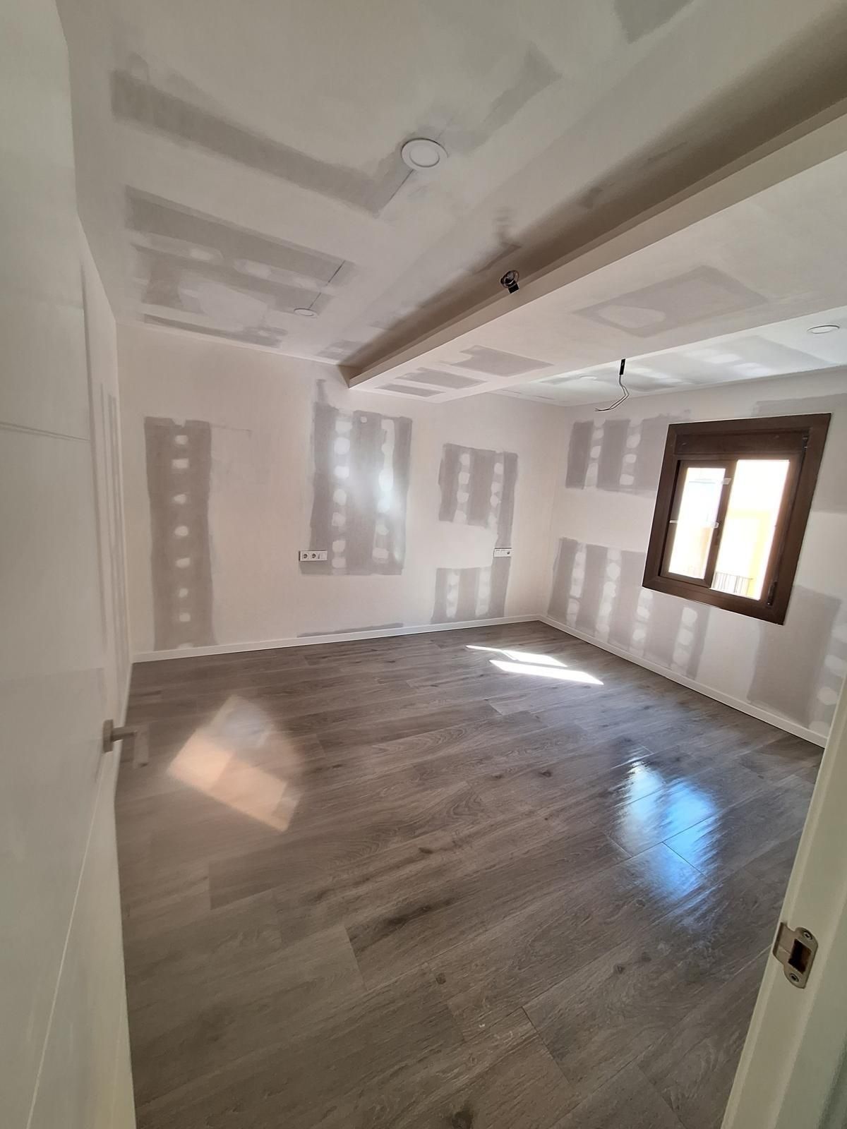 Habitación vacía sin amueblar con suelo de madera gris, paredes blancas y una ventana que deja entrar la luz del sol.