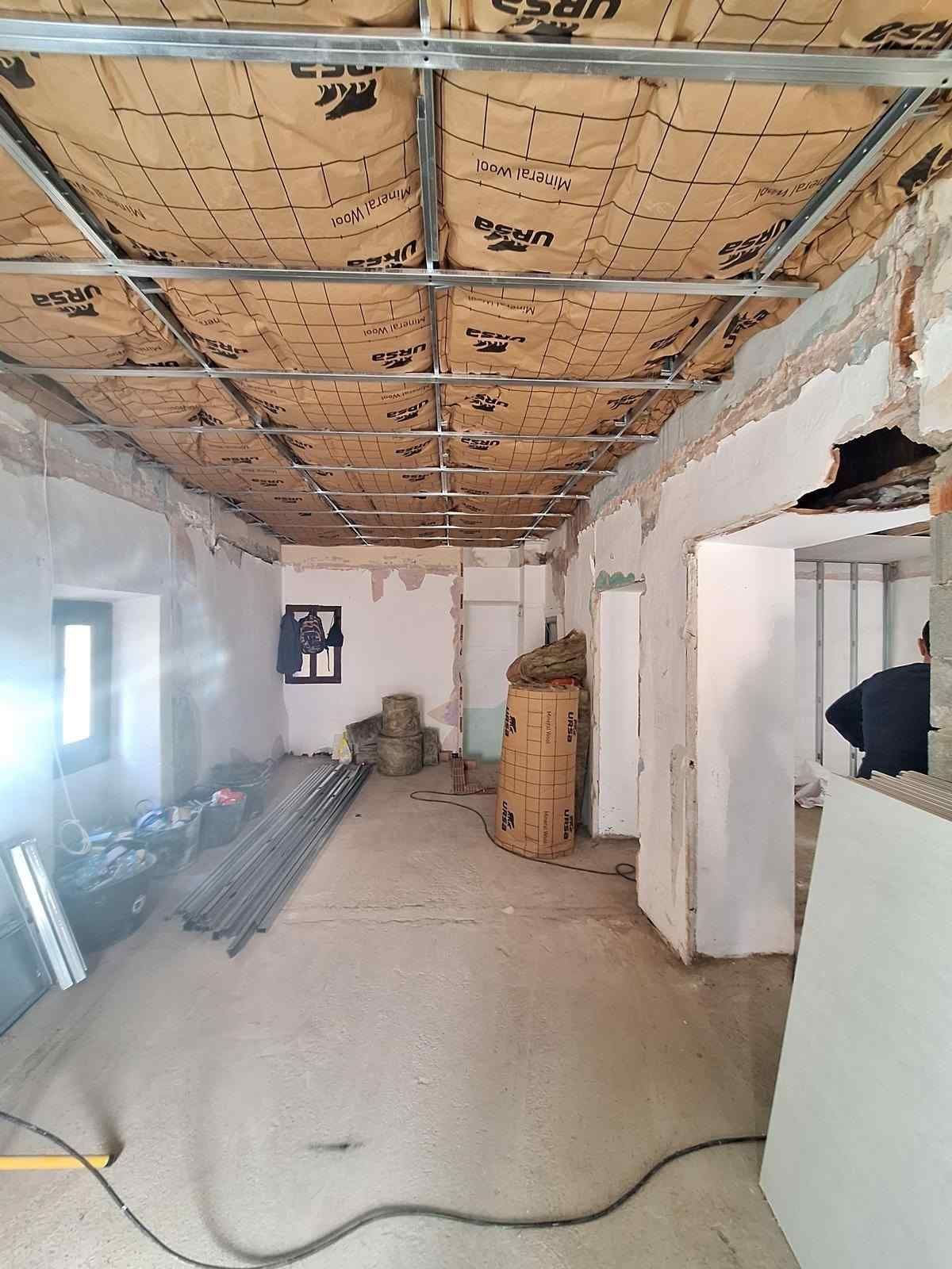 Habitación interior sin terminar con techo de aislamiento a la vista, paredes blancas y materiales de construcción esparcidos por el suelo.
