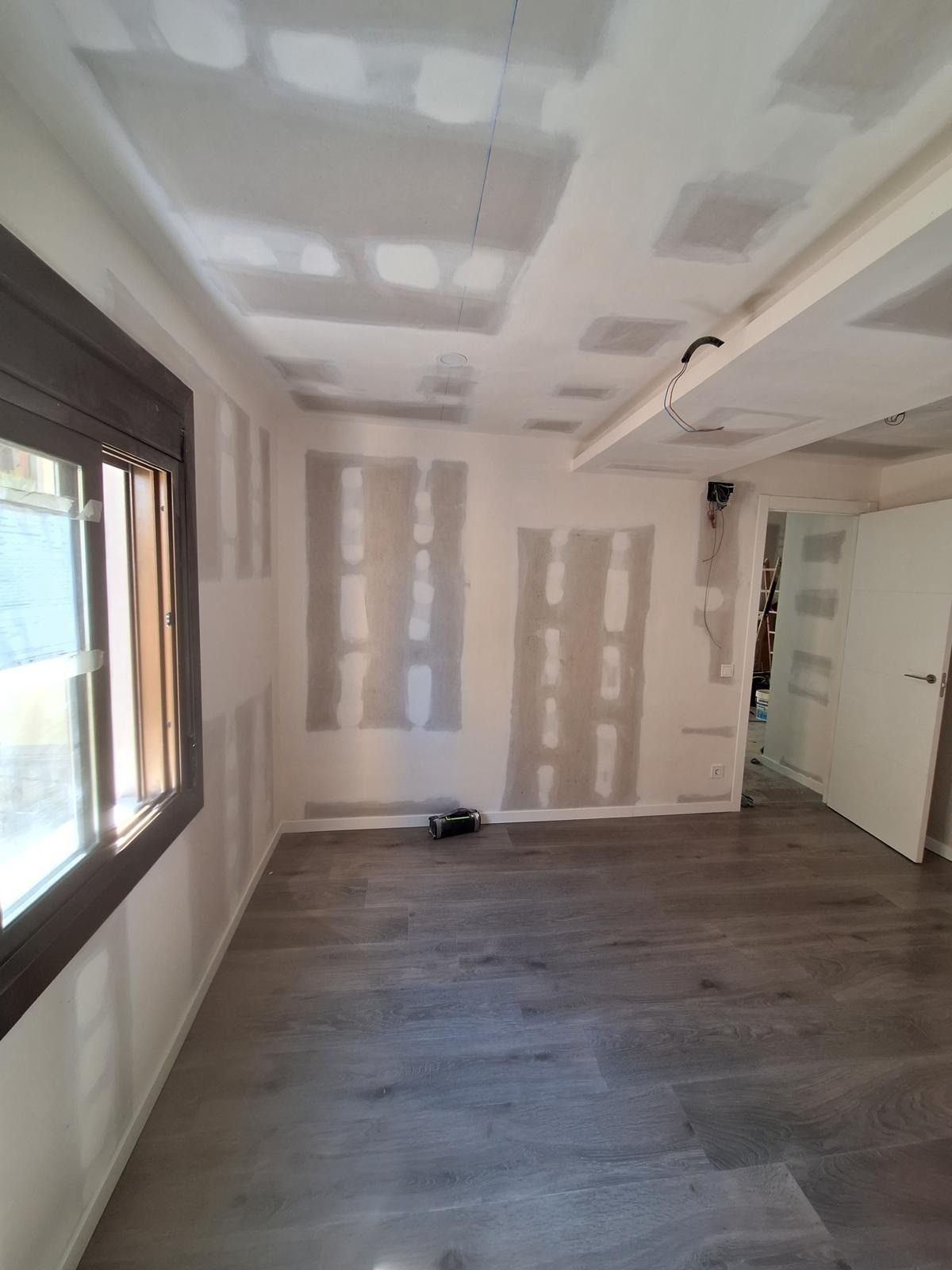 Habitación vacía reformada con paredes de pladur sin terminar, suelos de madera, ventana grande y puerta blanca.