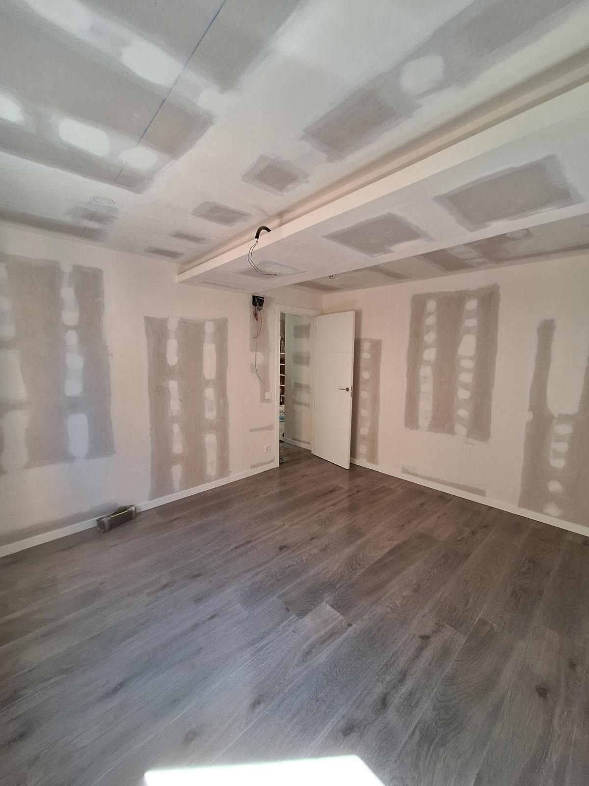 Habitación vacía con parches de yeso en las paredes, suelo de madera gris y una puerta abierta que da a un armario.