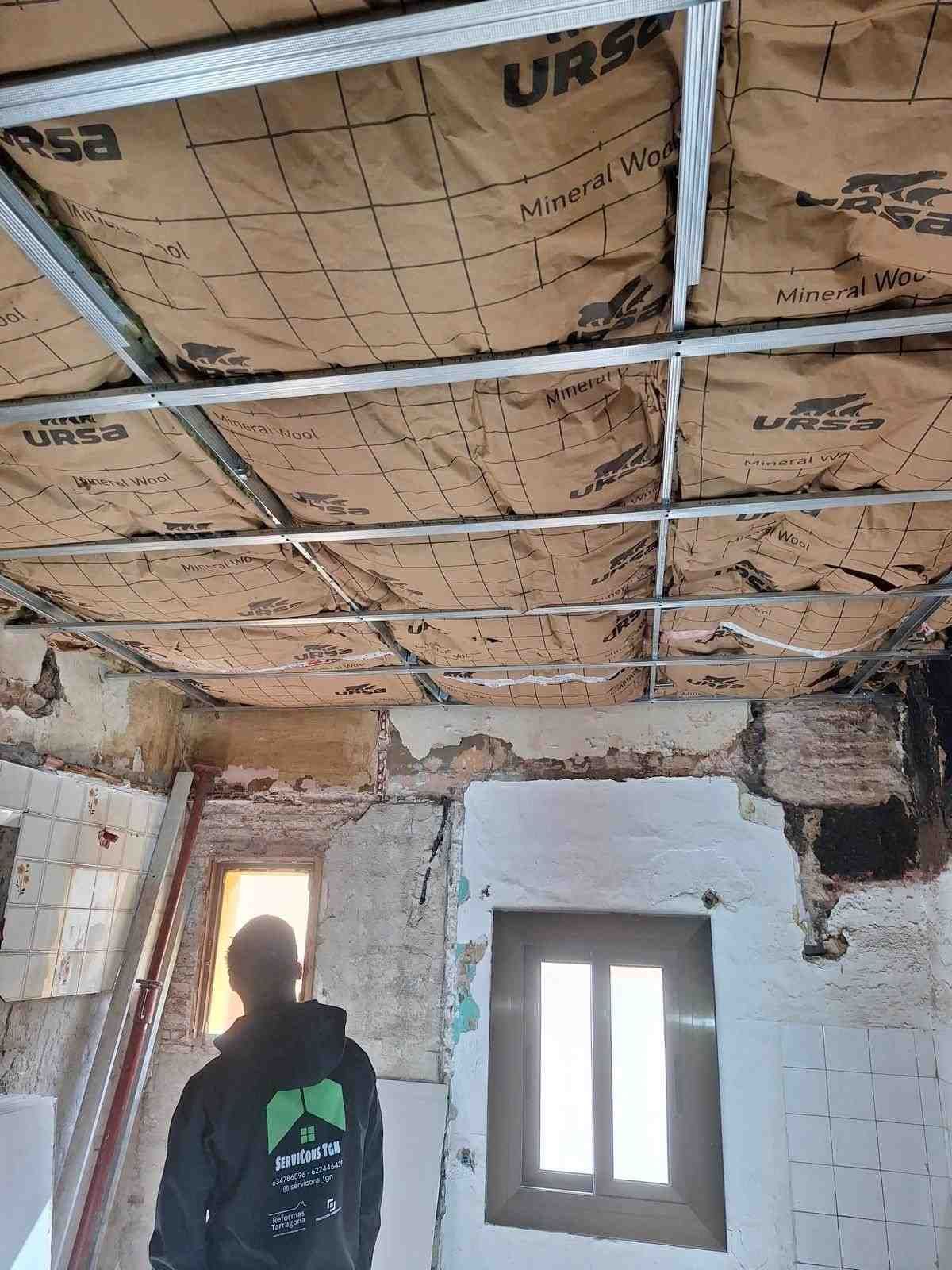 Habitación parcialmente renovada con aislamiento del techo a la vista, paredes dañadas y una persona de pie cerca de una ventana.