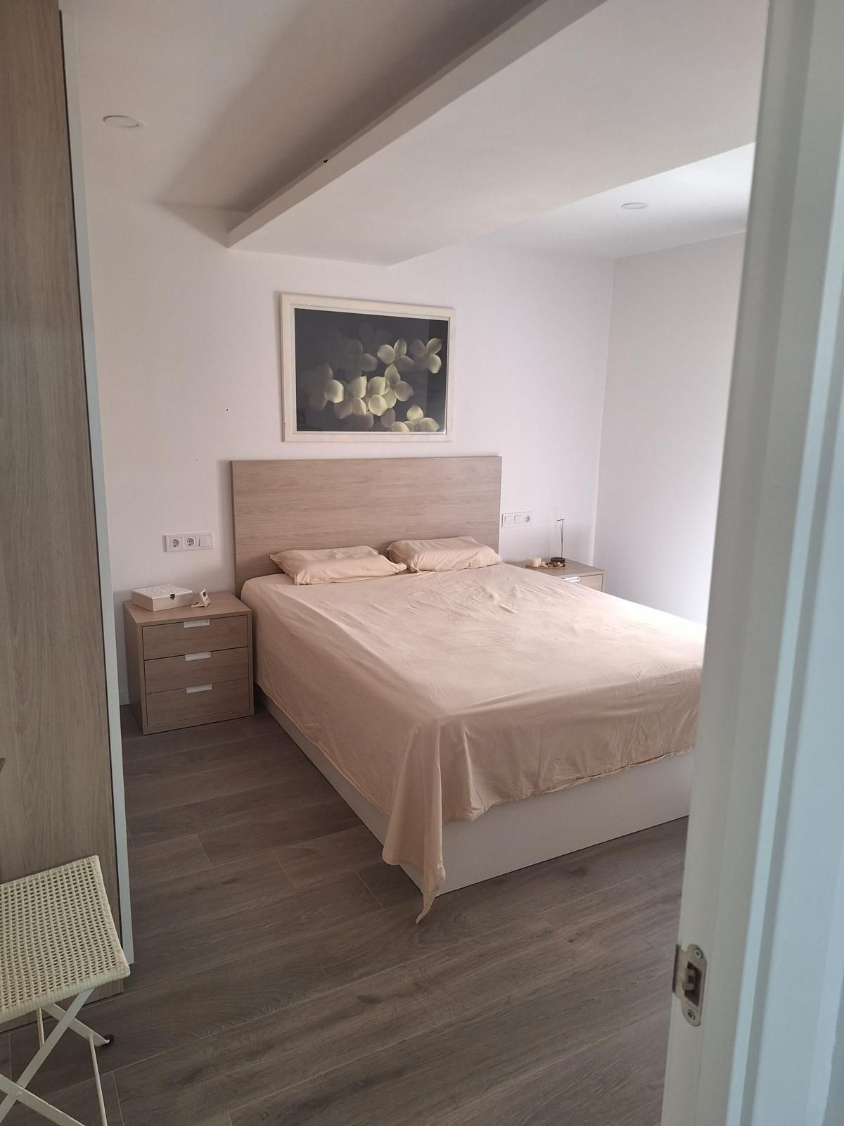 Habitación pequeña con cama doble, ropa de cama beige, mesita de noche de madera y cuadro enmarcado sobre el cabecero.