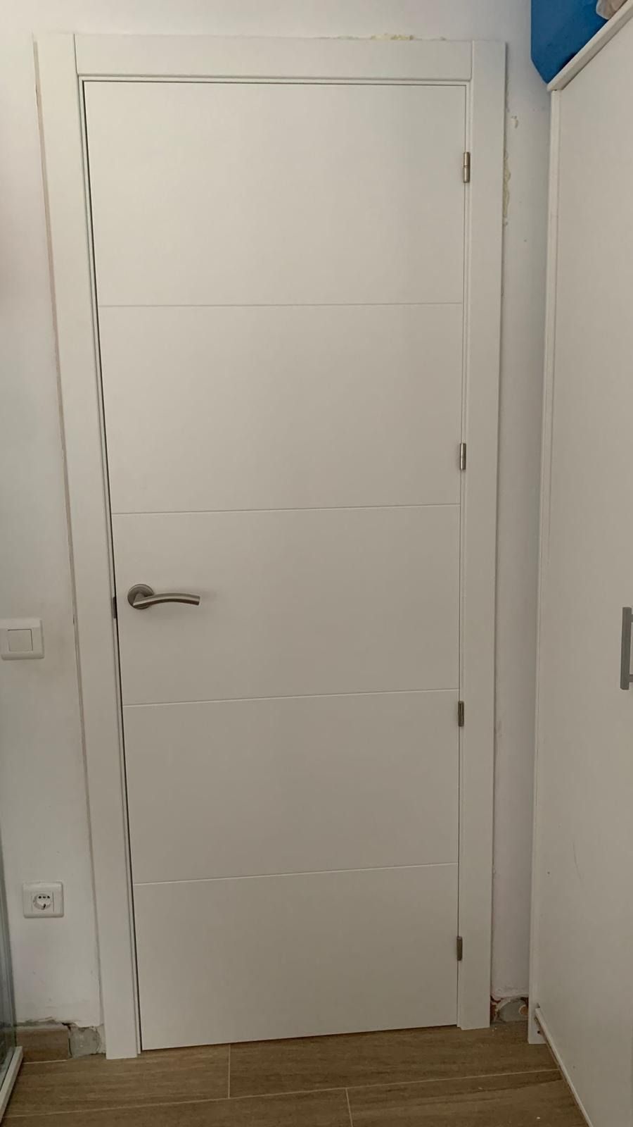 Puerta interior blanca con paneles horizontales y tirador plateado en un pasillo estrecho.