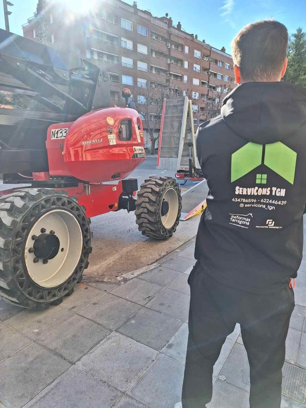 Una persona con una sudadera negra está de pie junto a un tractor rojo en la acera de la ciudad, bajo la luz del sol.