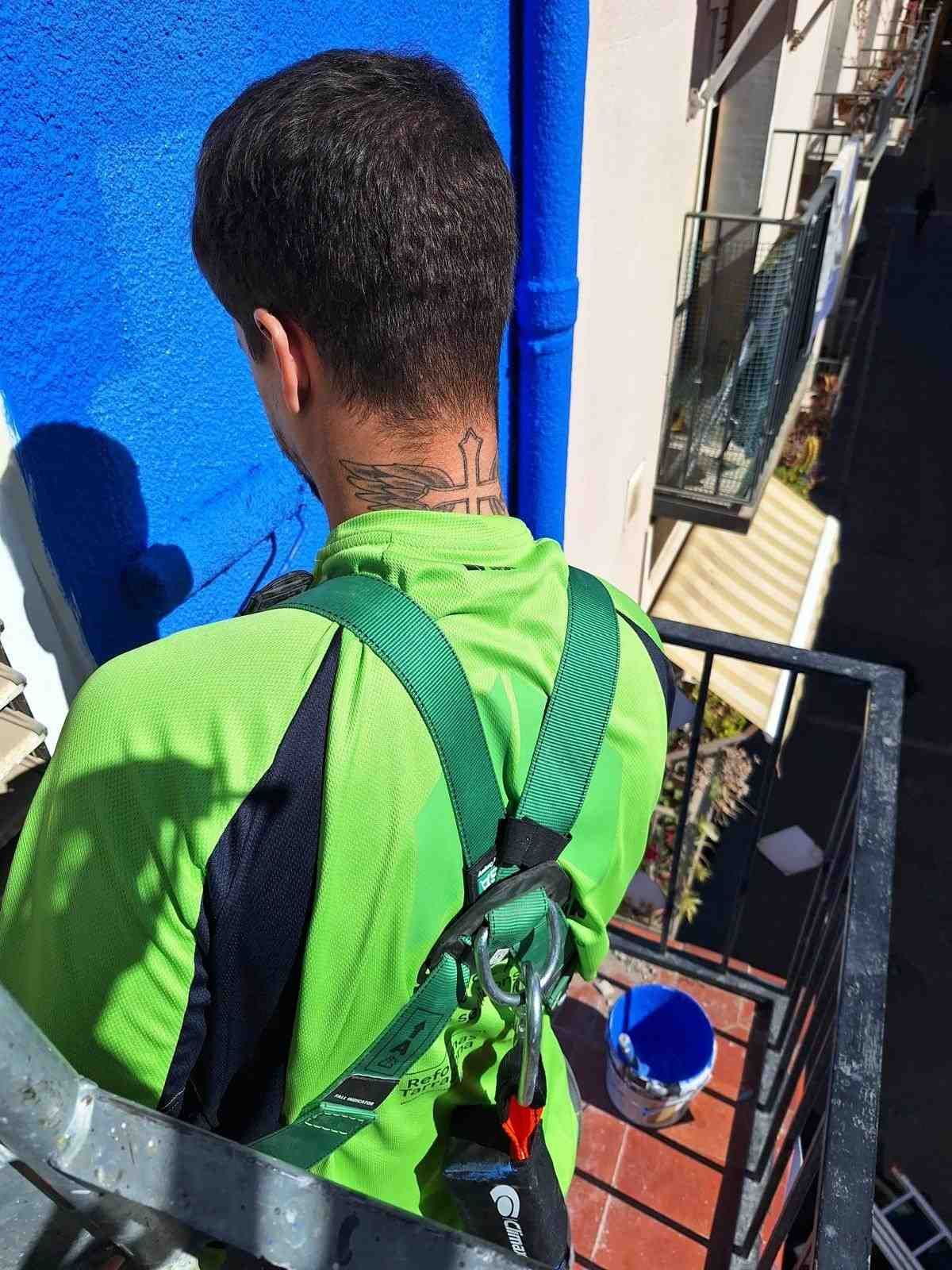 Un trabajador con una camisa verde brillante y arnés de seguridad está de pie en un balcón, mirando hacia una pared azul.
