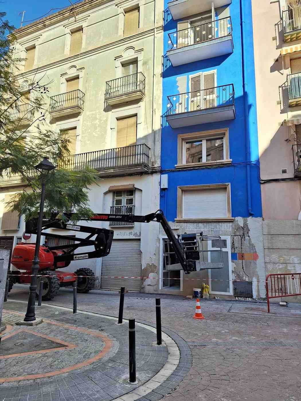 Un edificio de apartamentos azul con balcones y una grúa en la calle, situado en una esquina adoquinada.