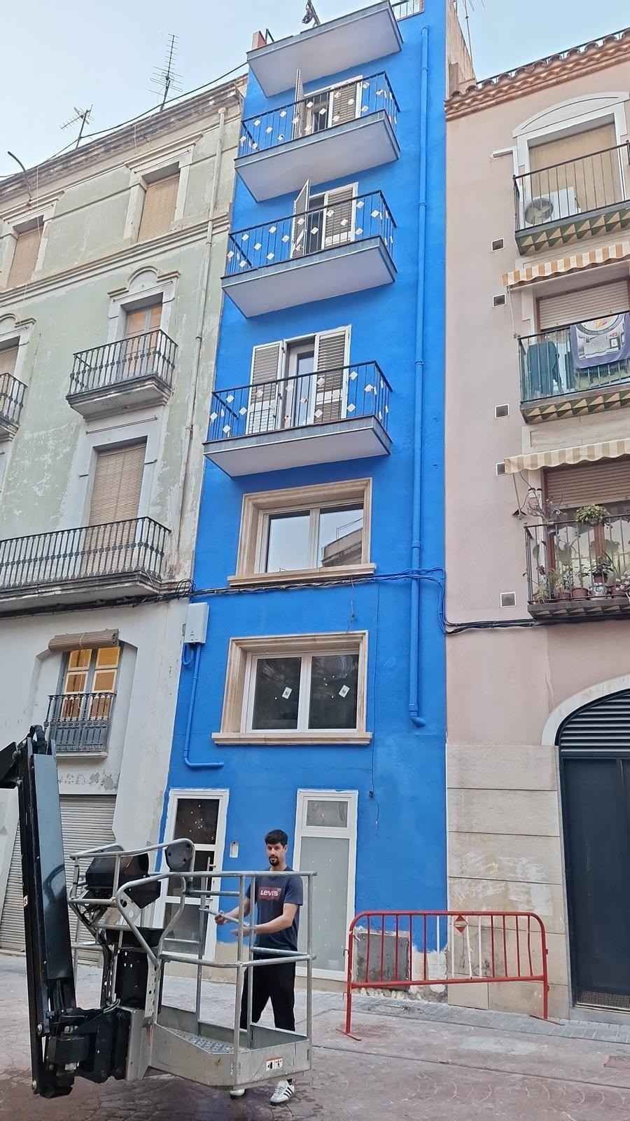 Edificio estrecho pintado de azul con balcones y ventanas en una calle de la ciudad, con una persona de pie debajo.