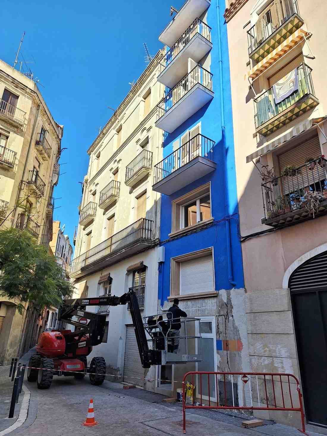 Trabajadores subidos a una plataforma elevadora pintando la fachada azul de un edificio de apartamentos en una calle estrecha, con un camión y barreras debajo.