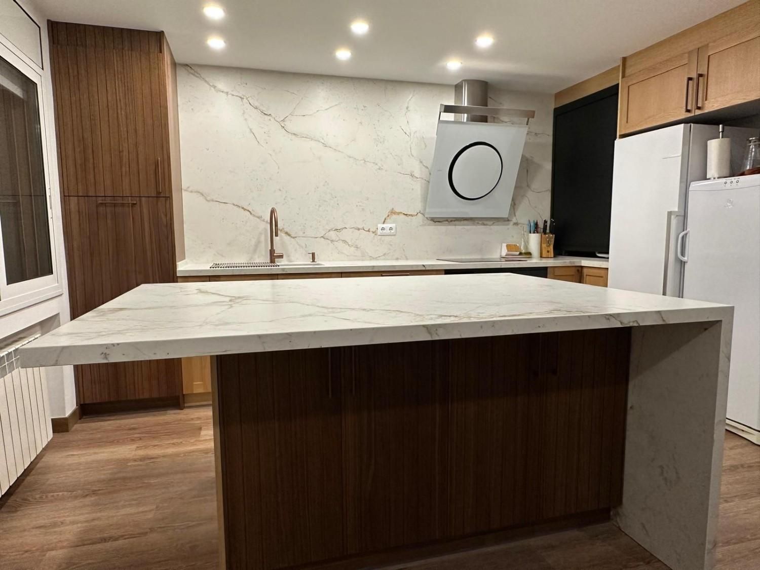 Cocina moderna con isla de mármol, gabinetes de madera, campana extractora de acero inoxidable e iluminación empotrada.