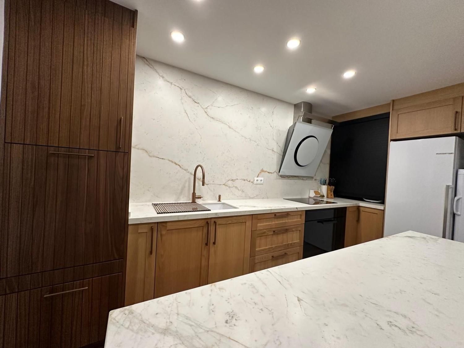 Cocina moderna con gabinetes de madera, encimeras de mármol, salpicadero blanco, fregadero y campana extractora de acero inoxidable.