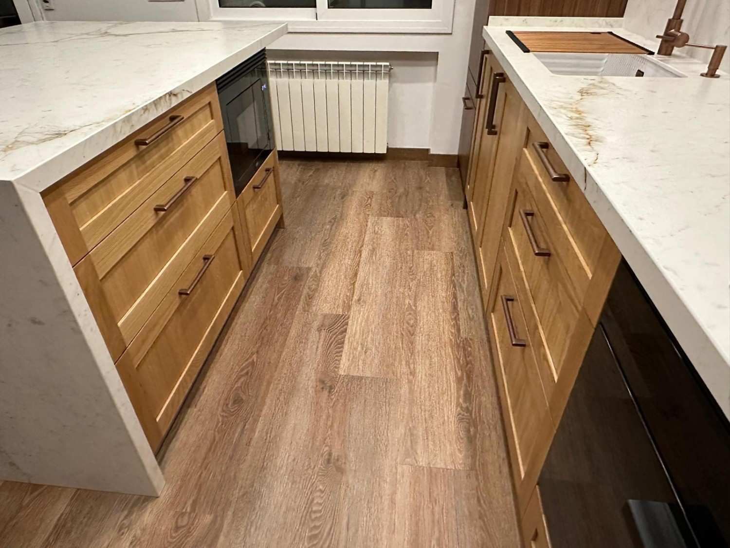 Cocina estrecha con encimeras blancas, armarios de madera y suelo de madera que da a una ventana y un radiador.