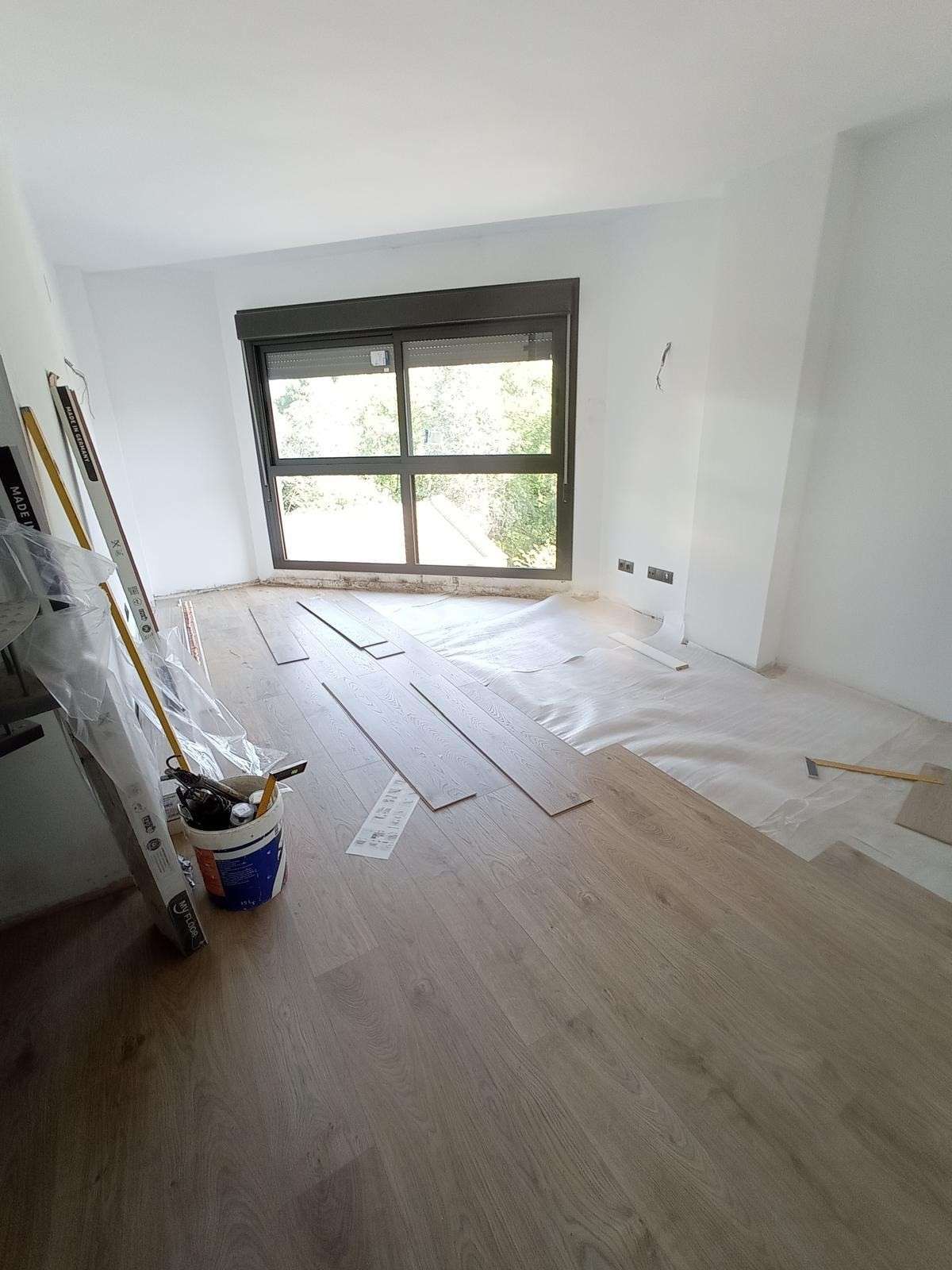 Habitación luminosa y vacía en proceso de renovación, con suelo de madera, materiales de pintura y una gran ventana.