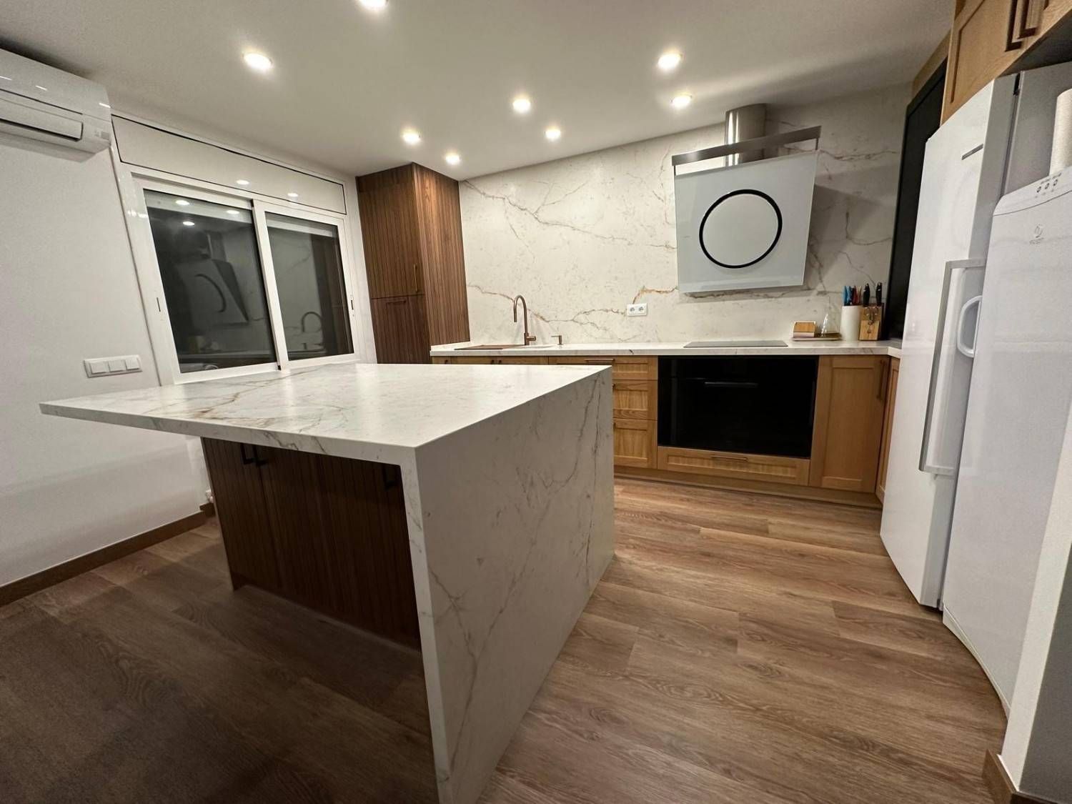 Cocina moderna con isla blanca, gabinetes de madera, encimeras de mármol y electrodomésticos de acero inoxidable.