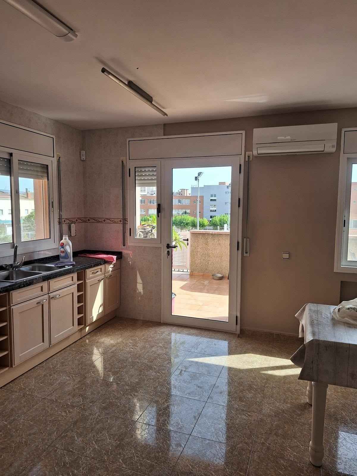 Cocina pequeña con armarios de color beige, suelo de baldosas, puerta que da a un patio soleado y una ventana sobre el fregadero.