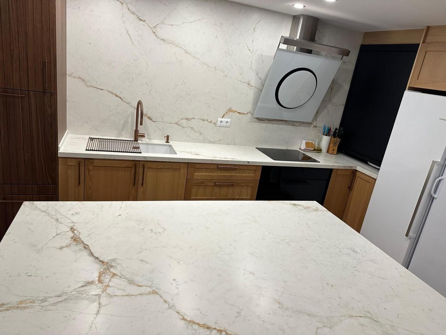 Cocina moderna con isla de mármol, armarios de madera, fregadero y electrodomésticos blancos.