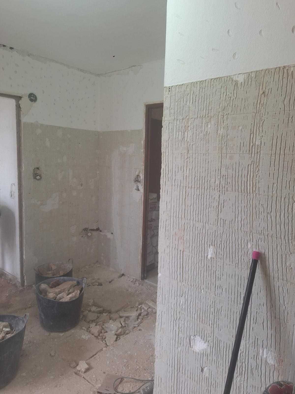 Habitación parcialmente renovada con paredes desnudas, escombros en el suelo y un cubo y herramientas cerca.