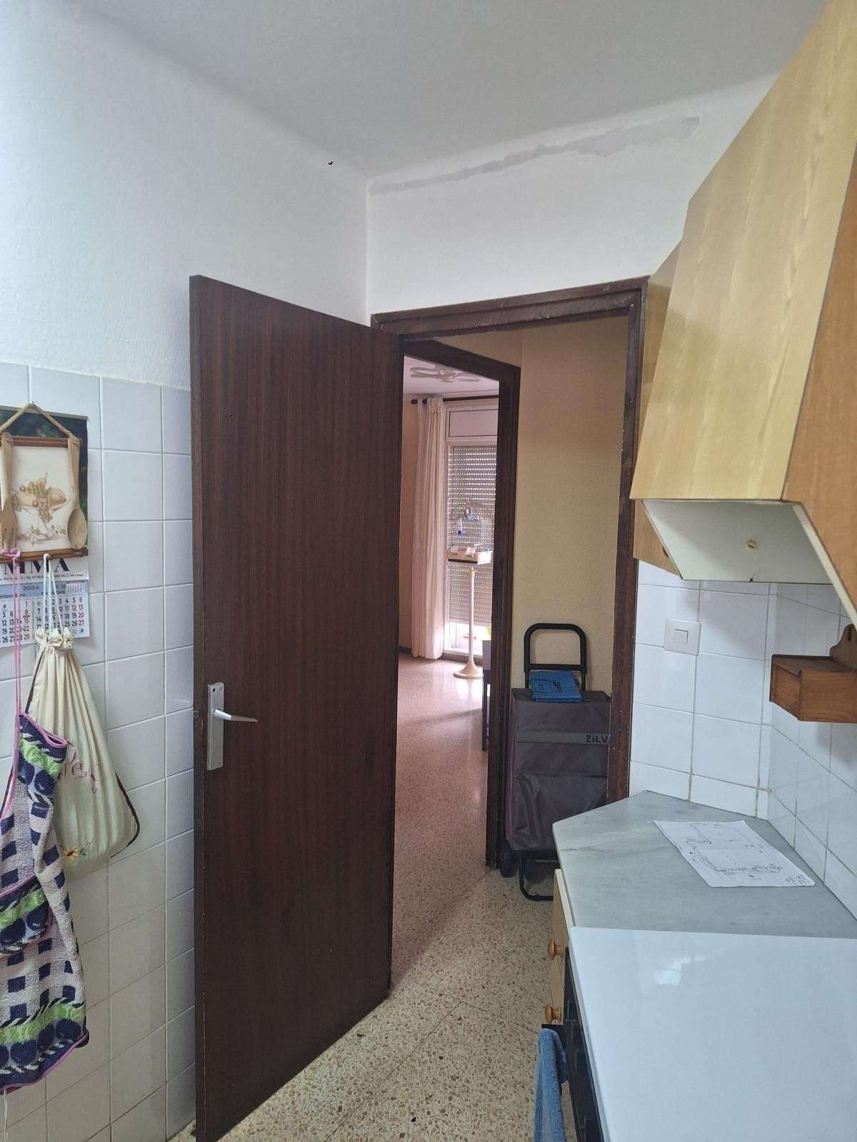 Estrecho pasillo de cocina con puertas marrones abiertas, armarios blancos y una bolsa sobre un carrito de mano en la entrada.