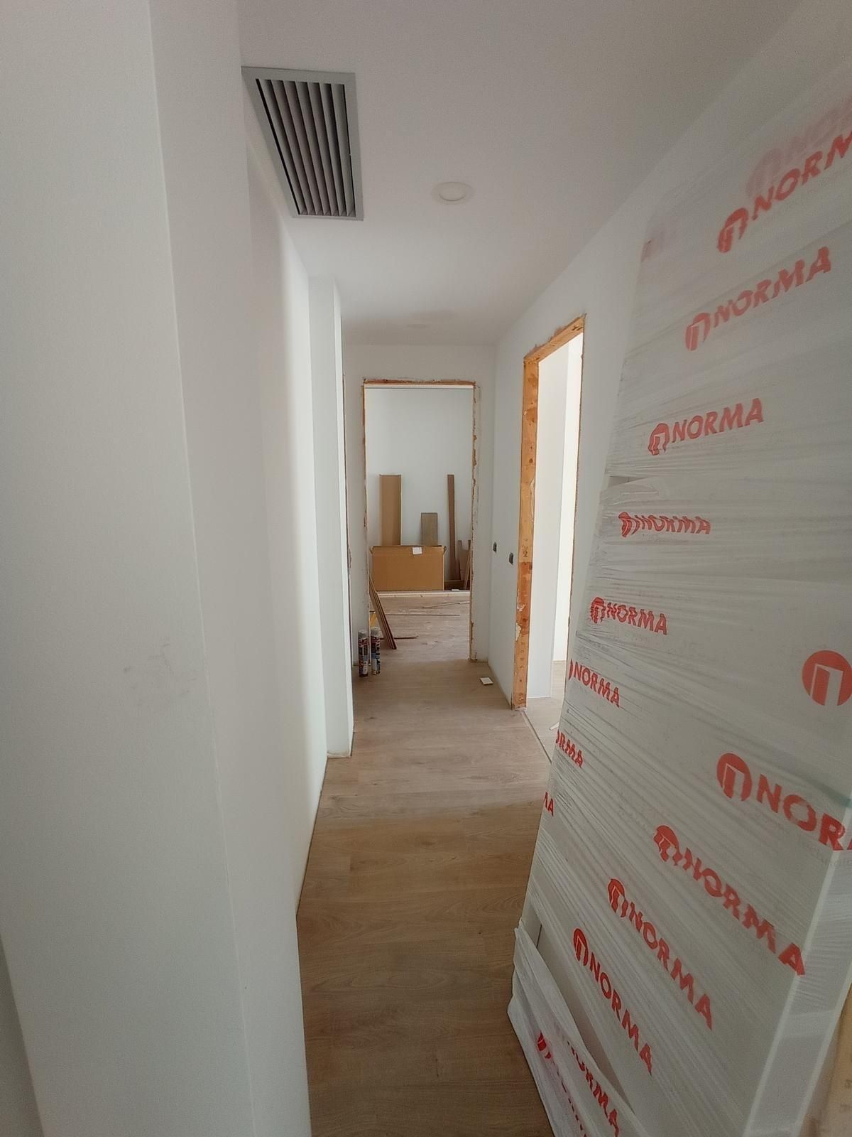 Pasillo estrecho en una casa en obras, con paredes blancas, suelo de madera y plástico protector en un lado.