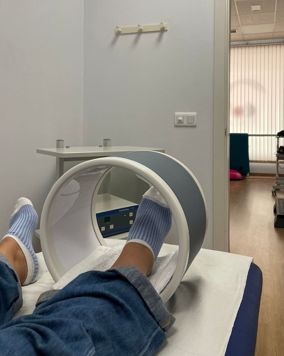 Persona que recibe terapia magnética, con los pies dentro de una bobina blanca y gris, en una mesa de masajes en un consultorio médico.