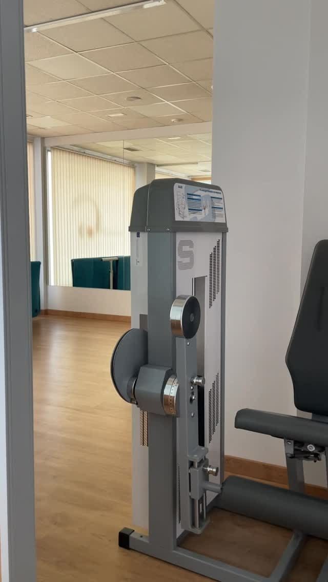 Aparatos de gimnasio en una sala luminosa con suelo de madera. Máquina de pesas gris enfocada. Otra sala visible al fondo.