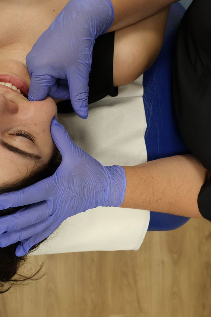 Manos enguantadas realizando un masaje facial a una persona tumbada en una camilla de tratamiento.