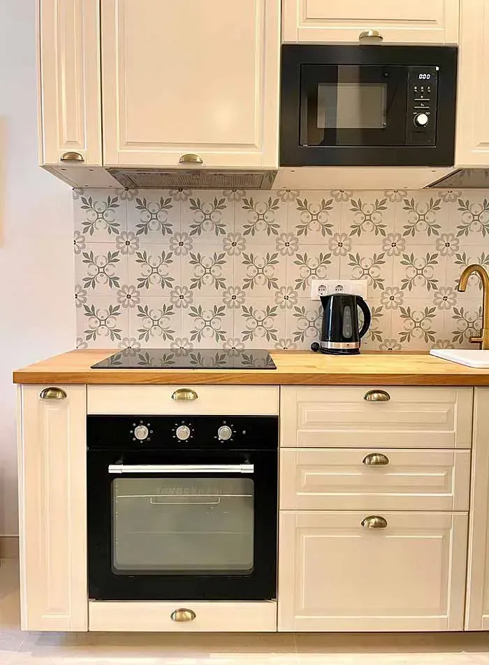 Una cocina con gabinetes blancos, un horno negro, un microondas, una tetera y un fregadero.