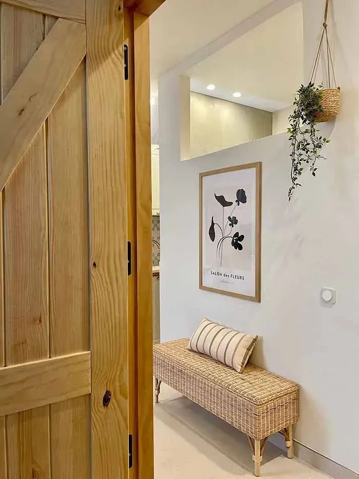 Una puerta de madera se abre a un pasillo con un banco y un cuadro en la pared.