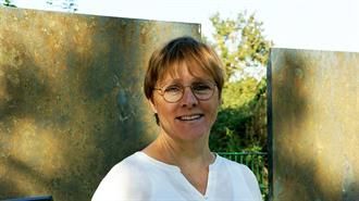 Eine Frau mit Brille und weißem Hemd steht vor einer Wand.