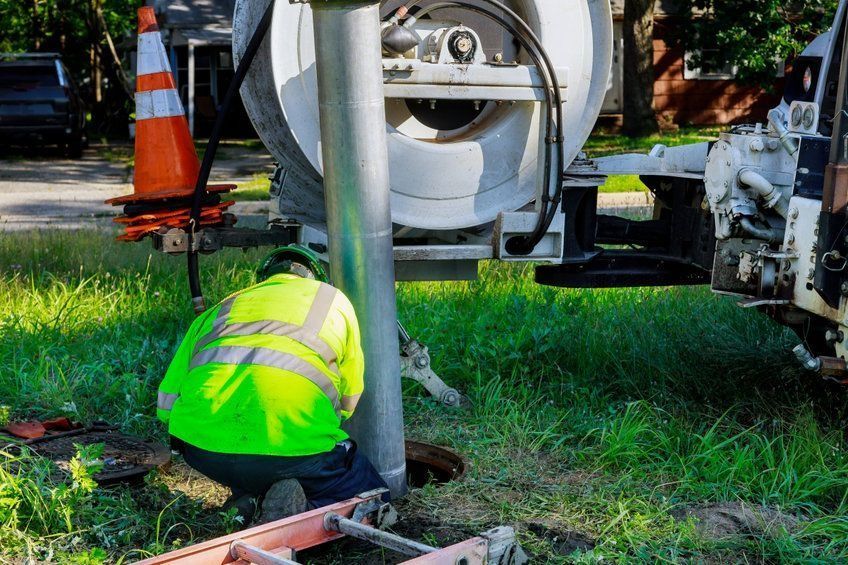 Un trabajador con un chaleco reflectante se agacha junto a un equipo de servicios públicos cerca de un camión en un terreno cubierto de hierba.