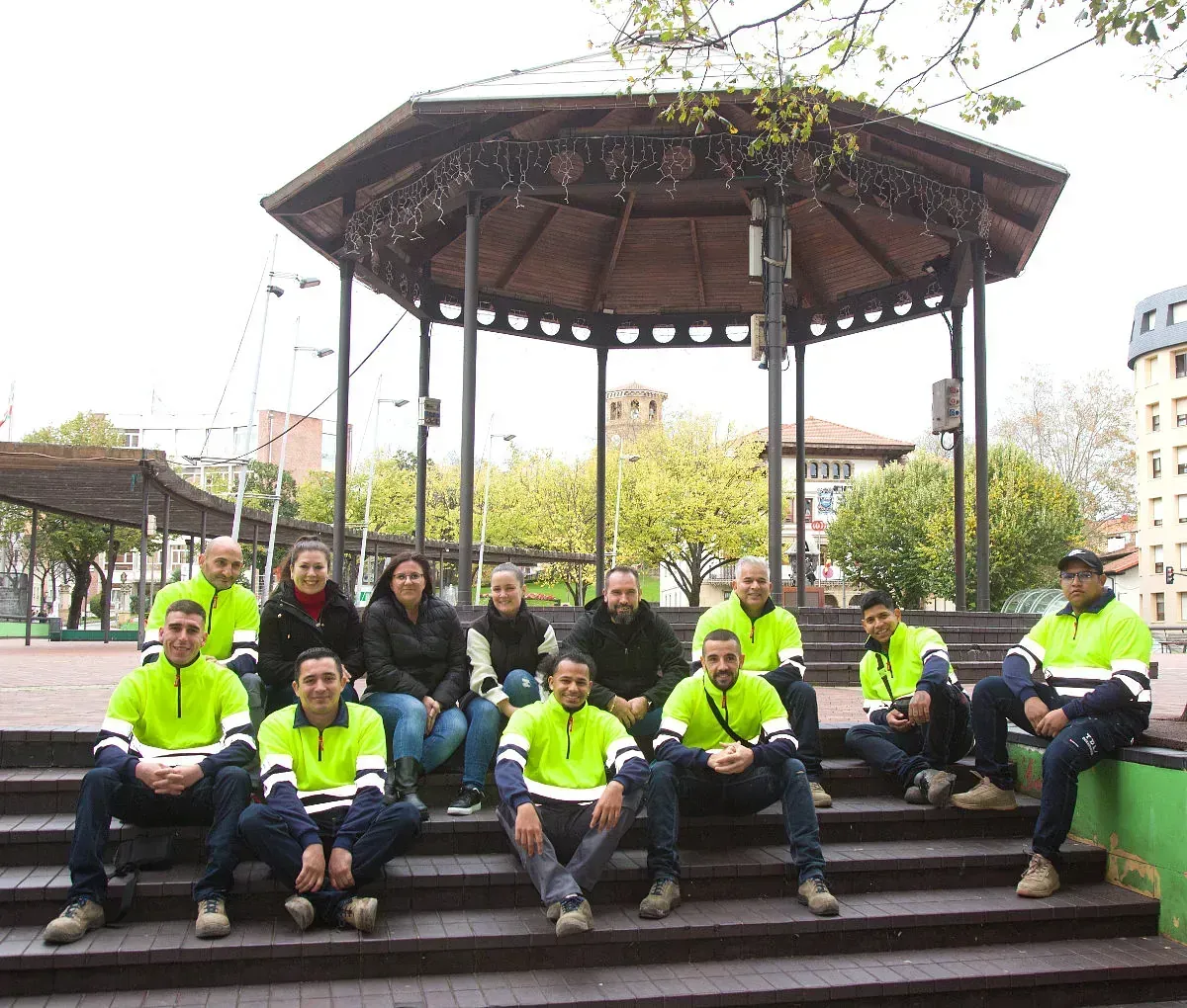 Un grupo de personas con chaquetas de colores neón están sentadas en unas escaleras bajo una glorieta en un parque.