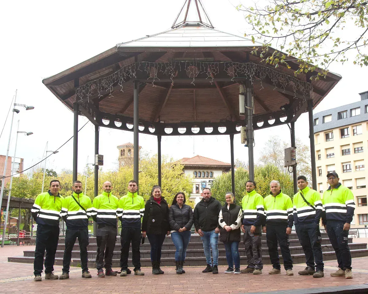 Un grupo de personas con chaquetas de colores neón posan frente a una glorieta en una plaza de la ciudad.