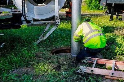 Un trabajador con una chaqueta de color neón se agacha junto a un poste al lado de un camión, inspeccionando un agujero en el suelo.