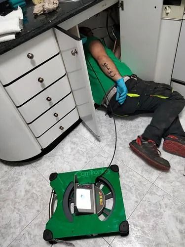 Un trabajador con camisa verde y guantes azules se encuentra debajo de un escritorio, con un dispositivo de inspección verde en el suelo.